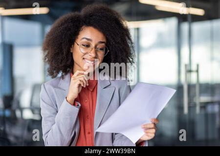 Una giovane donna professionale con capelli ricci che indossa occhiali recensisce i documenti con un'espressione premurosa in un moderno ambiente d'ufficio. Foto Stock