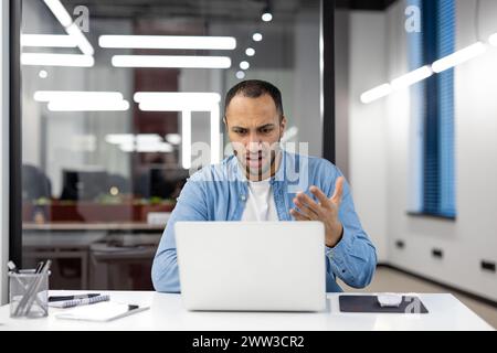 Giovane professionista di sesso maschile in un ufficio moderno che sembra stressato e confuso mentre lavora al suo notebook, mostrando un segno di sfide aziendali. Foto Stock