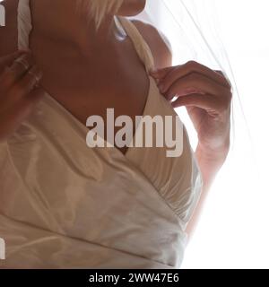 Questa fotografia intima si concentra sulla silhouette di una sposa, catturando la grazia e il fermento mentre regola il suo abito. La morbida retroilluminazione crea un delicato gioco di luci e ombre, mettendo in risalto i dettagli raffinati del suo abbigliamento da sposa. Eleganza nuziale: Uno sguardo di grazia nell'abbigliamento da sposa. Foto di alta qualità Foto Stock