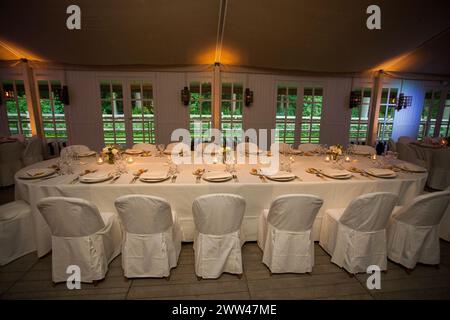 Una sofisticata sala banchetti, pronta per gli ospiti, con tavoli vestiti con immacolate lenzuola bianche e adornati con delicate composizioni floreali. L'illuminazione intima diffonde un caldo bagliore, completando la serena vista esterna visibile attraverso le finestre. Elegante disposizione per banchetti con biancheria bianca immacolata e illuminazione intima. Foto di alta qualità Foto Stock