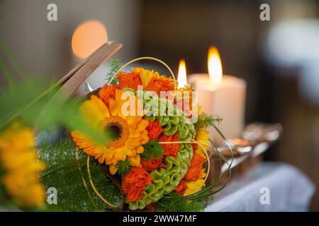 Questa immagine si concentra su un pezzo centrale floreale, caratterizzato da fiori di arancio brillante e da un'intricata vegetazione, completata dal bagliore ambientale delle candele vicine. L'illuminazione soffusa esalta i colori naturali e crea un ambiente accogliente. Un caldo bagliore di candelabro che accentua un centrotavola floreale. Foto di alta qualità Foto Stock