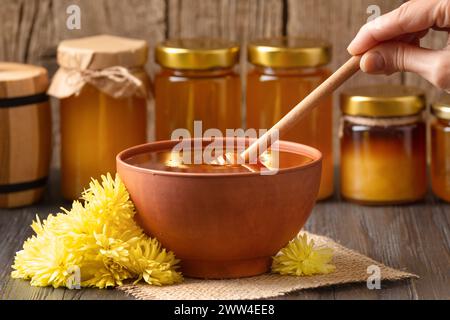 Una mano abbassa un cucchiaio di legno in una ciotola di ceramica con miele sullo sfondo di vari vasetti di miele. Cucchiaio di miele in un recipiente di argilla con fiore Foto Stock
