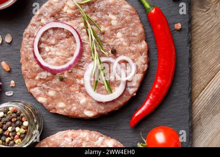 Hamburger crudo di manzo macinato, rosmarino, verdure e spezie su un piatto di pietra nera su uno sfondo di legno, vista dall'alto, primo piano. Foto Stock