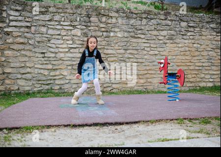 Bambina attiva, studentessa di età elementare gioca a hopscotch, a turno salta su quadrati segnati a terra. Giochi per bambini di strada Foto Stock