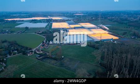 Duffel, Belgio, 20 marzo 2024, una prospettiva aerea durante il crepuscolo mostra ampie serre industriali, le loro luci si illuminano come fari nel paesaggio rurale. Il contrasto tra ambienti artificiali e naturali è sorprendente, mentre il giorno sbiadisce fino alla notte. Crepuscolo luminoso sulle serre industriali nel paesaggio rurale. Foto di alta qualità Foto Stock