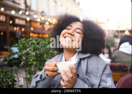 Moda Street style ritratto di attraente giovane bellezza naturale African American donna con capelli afro in tweed giacca posa all'aperto in sidewalk cafe. Signora felice con la tazza di caffè nella grande città Foto Stock