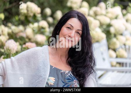 Una donna sorridente con i capelli neri, con un abito floreale e uno scialle bianco, siede su una panchina del parco circondata Foto Stock