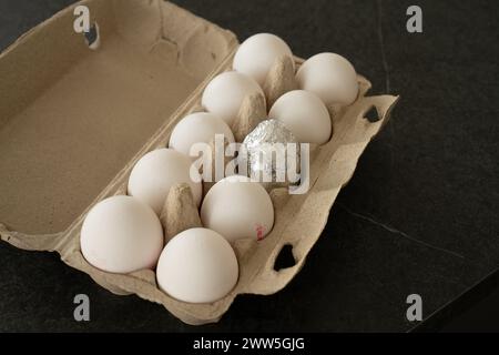 Mettere la scatola delle uova aperta su un tavolo scuro con dieci uova bianche. Un uovo al centro è avvolto in un foglio d'argento, come uovo di pasqua. Foto Stock