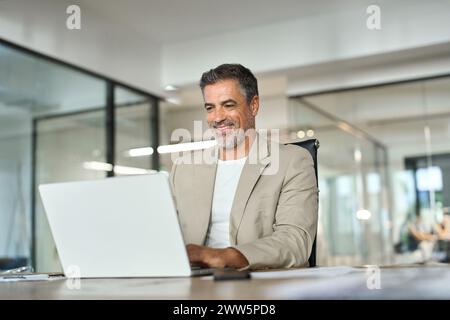 Un manager di banca maturo e felice che guarda un notebook in ufficio seduto alla scrivania. Foto Stock