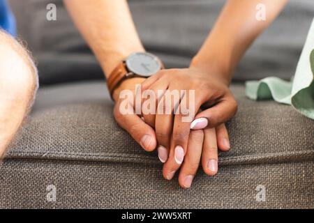 Una coppia eterogenea si tiene per mano, mostrando unità e affetto a casa sul divano Foto Stock