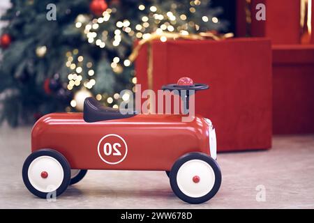 Vista dell'auto retrò rossa per bambini sullo sfondo di regali in scatole rosse con archi dorati, ghirlanda decorata con alberi di Natale, luci a corde, palline rosse e blu. Sfondo, spazio copia, bokeh. Foto Stock