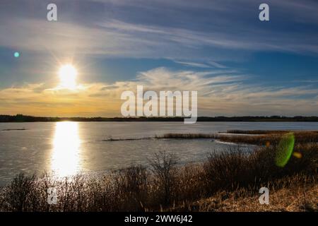 Calmo tramonto serale invernale vicino alla riva di un lago ghiacciato con cespugli, prati, riflessi e un bel cielo sullo sfondo Foto Stock