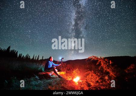 Giovane uomo che scatta foto di cielo stellato in montagna. Fotografo maschio seduto su una macchina fotografica di legno e messa a fuoco. Uomo con macchina fotografica seduta accanto al fuoco del campo. Foto Stock