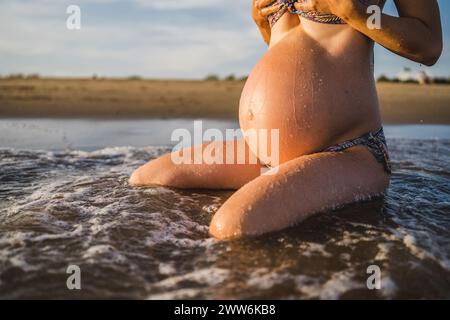 donna incinta seduta sulla riva del mare, bagnata dalle onde, dalle gocce e dalla luce serale Foto Stock