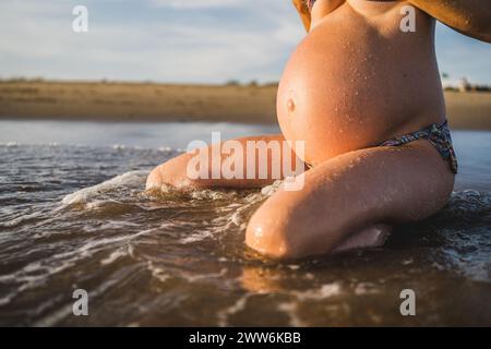 donna incinta seduta sulla riva del mare, bagnata dalle onde, dalle gocce e dalla luce serale Foto Stock