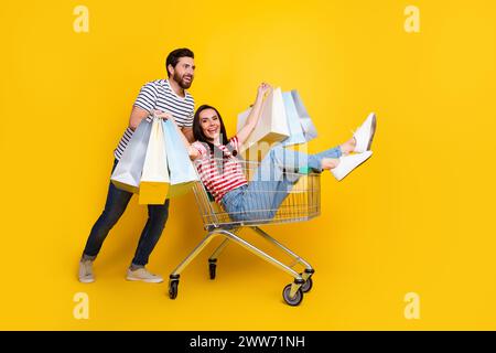 Foto a grandezza naturale di un uomo con abiti a righe positivi cavalca una ragazza in un carrello con borse della spesa in braccio isolate su sfondo di colore giallo Foto Stock