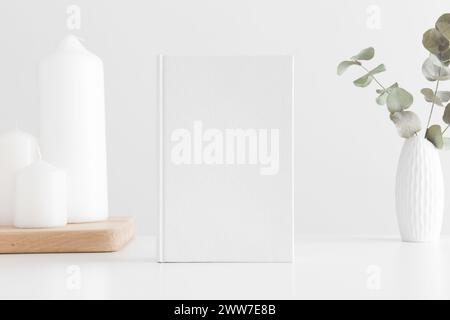 Modello di libro bianco con eucalipto in pentola e candele su un tavolo bianco. Foto Stock