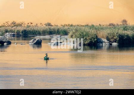 Pescatore in barca a remi sul fiume Nilo al sole, paesaggio panoramico con rocce nell'acqua e riflessi dune di sabbia ad Assuan, Egitto Foto Stock