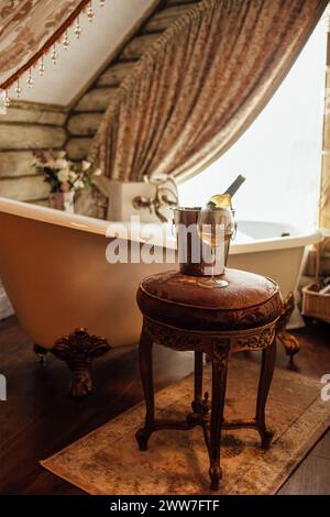 Primo piano di un bicchiere di vino e di una bottiglia in un secchio di ghiaccio su un'elegante sedia in bagno. Un bagnoschiuma pieno accanto alla finestra. Interi vintage Foto Stock