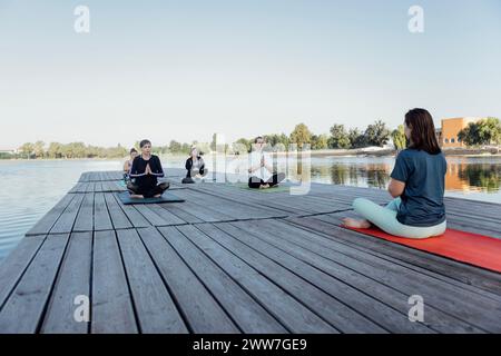 Lezione di yoga all'aperto con istruttore vicino al fiume. Le giovani donne siedono a gambe incrociate sui tappetini e meditano su un molo di legno. Un gruppo di donne multietniche Foto Stock