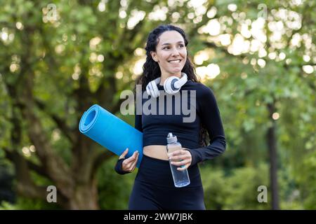 Appassionato di fitness sorridente che porta con sé un tappetino da yoga e una bottiglia d'acqua, preparandosi per una sessione di allenamento all'aperto circondata da alberi. Foto Stock