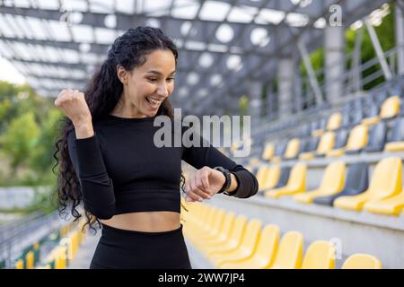 Donna incentrata sul fitness in abbigliamento sportivo che utilizza uno smartwatch in uno stadio sportivo, incarnando salute e stile di vita attivo. Foto Stock