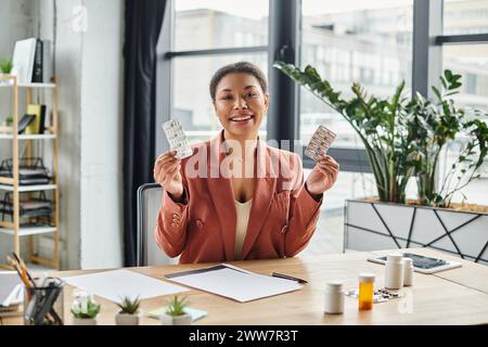 gioiosa nutrizionista afroamericana che mostra diversi farmaci mentre siede nel suo ufficio Foto Stock