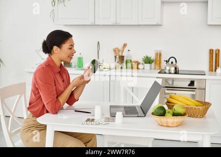nutrizionista afroamericano felice che tiene in mano avocado maturo e dà consigli sulla dieta sul portatile in cucina Foto Stock