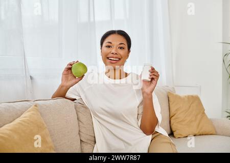 felice dietologo afroamericano che tiene in mano mela verde e integratori e sorride nel soggiorno Foto Stock