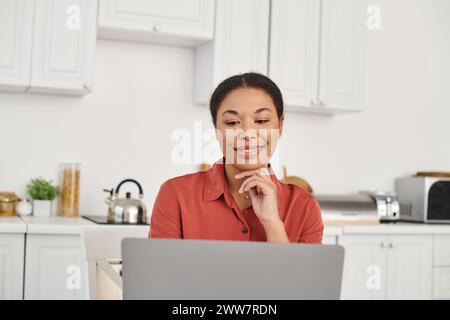 Lavoro a distanza di allegra giovane nutrizionista afroamericana che usa un computer portatile nella sua cucina, donna Foto Stock