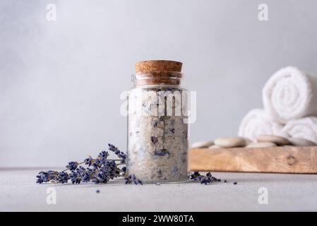 Un ambiente spa sereno con un vaso di vetro di sale aromatico di lavanda sigillato con un sughero, circondato da fiori di lavanda sparsi, asciugamani bianchi e liscio Foto Stock