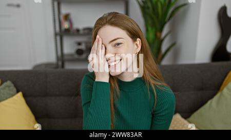 Una giovane e allegra donna caucasica copre metà del suo viso giocosamente in un salotto accogliente ed elegante. Foto Stock