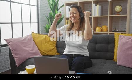 Una donna ispanica eccitata con le cuffie utilizza un computer portatile in un accogliente ambiente domestico, esprimendo gioia. Foto Stock