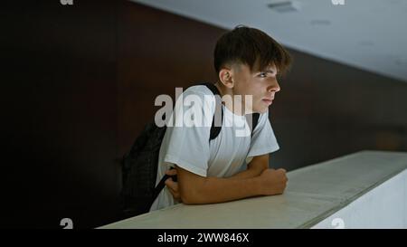 Un giovane adolescente caucasico contemplativo con uno zaino si appoggia su un bancone in un ambiente scolastico. Foto Stock