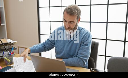 Bell'ispanico di mezza età che indossa un maglione e una cravatta che lavora alla scrivania del suo ufficio su un notebook. Foto Stock