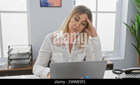 Donna caucasica stressata che soffre di mal di testa in un ufficio moderno, concentrandosi sul suo lavoro su un notebook. Foto Stock