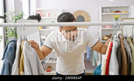 Uomo ispanico che esamina i vestiti in un luminoso guardaroba interno, riflettendo sulle scelte di moda. Foto Stock