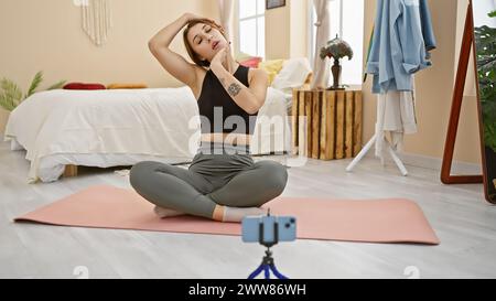 Una giovane donna filma una sessione di yoga con uno smartphone su un treppiede nella sua accogliente camera da letto. Foto Stock