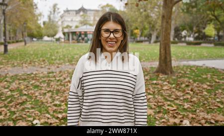 Una giovane donna ispanica sorridente si trova in un parco autunnale con occhiali e un maglione a righe. Foto Stock
