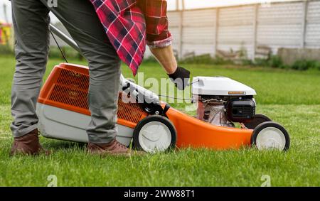 Un giardiniere irriconoscibile con guanti protettivi avvia il rasaerba prima di tagliare il prato verde nel suo cortile. L'uomo con rasaerba motorizzato si prende cura di prati paesaggistici. Foto Stock