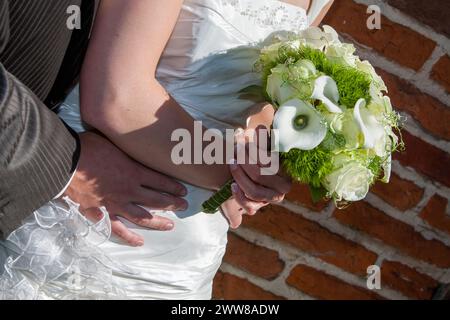 L'immagine incornicia splendidamente le mani agitate di una sposa e di uno sposo sullo sfondo del suo elegante bouquet nuziale e della texture senza tempo di una parete in mattoni, catturando un momento di unità e impegno nella luce dorata del sole. Abbraccio delle promesse: Gli sposi si stringono la mano con il bouquet nuziale. Foto di alta qualità Foto Stock