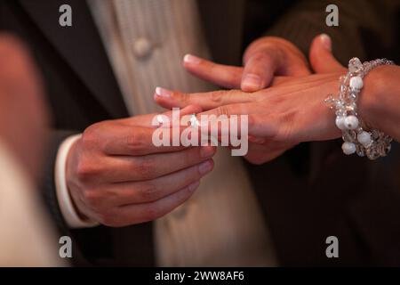 Questa fotografia cattura un momento toccante all'interno di una cerimonia nuziale: Il delicato posizionamento di una banda nuziale sul dito della sposa. L'attenzione alle mani sullo sfondo morbido evoca l'intimità e il significato del legame matrimoniale. Voti scambiati: Il momento tenero del posizionamento dell'anello durante una cerimonia nuziale. Foto di alta qualità Foto Stock