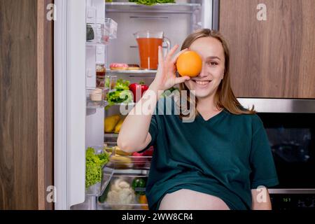 Giovane donna incinta con un'arancia vicino al frigorifero di casa. Mangiare sano. Foto Stock
