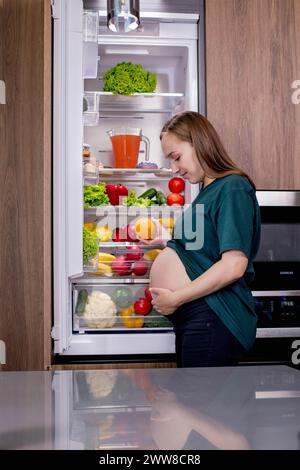 Giovane donna incinta con un'arancia vicino al frigorifero di casa. Mangiare sano. Foto Stock