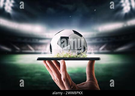 Concetto di trasmissione online in diretta del campionato di calcio con la mano che tiene in mano un telefono cellulare con palla e stadio di calcio sullo sfondo. Foto Stock