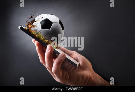 Concetto di guardare il calcio online con la mano che tiene in mano un telefono cellulare con palla e sfondo isolato grigio scuro. Foto Stock