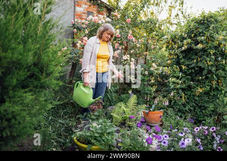 Un'anziana donna sorridente annaffia i fiori in vasi appesi da una lattina da innaffiare. Una donna anziana positiva in abiti casual si prende cura delle piante. Un bell'fu Foto Stock