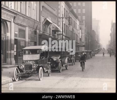 Vista sulla strada dell'ottava strada. Una fila di automobili fiancheggia il lato sinistro della strada di fronte a una fila di edifici commerciali. Un uomo corre in bicicletta per il centro della strada. Intorno al 1900 St-Louis, Missouri. STATI UNITI Foto Stock