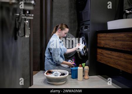 Donna seduta di fronte a una lavatrice che carica abiti sporchi, tovaglioli per lavare abiti colorati. Foto Stock