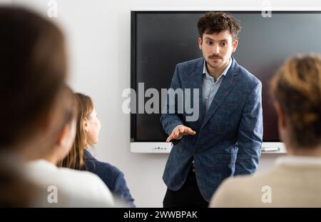 Istruttore maschile esperto e positivo che spiega l'argomento a un gruppo di persone durante la lezione di lingua straniera in udito Foto Stock